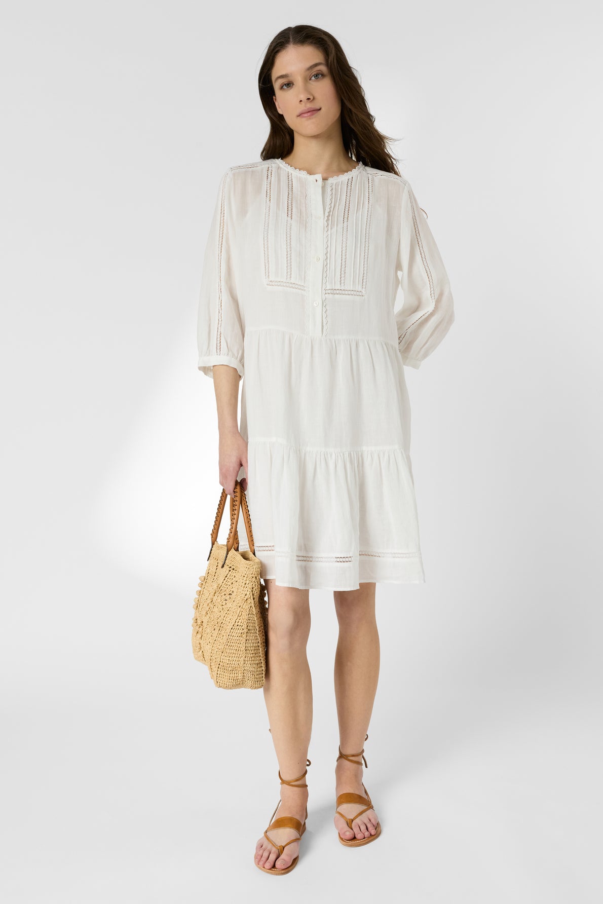 Woman in a loose, white, knee-length dress with long sleeves holds a woven straw bag and wears tan sandals, standing against a plain light background.