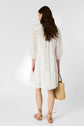 A woman with long brown hair, wearing a white, knee-length dress and brown sandals, stands facing away, holding a woven straw bag in her left hand against a plain white background.