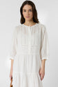 Woman with long brown hair wearing a white, long-sleeve, buttoned dress with vertical lace details, standing against a plain light background, looking directly at the camera with a neutral expression.