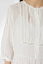 A close-up of a person wearing a white, long-sleeved blouse with lace trim and vertical embroidered details on the front. Only the torso and part of one arm are visible.