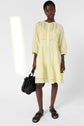 A woman wearing a light yellow, knee-length dress with three-quarter sleeves and black sandals holds a large black woven handbag. She also wears a gold ring and stands against a plain white background.