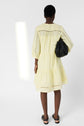 A person stands facing away, wearing a light yellow, knee-length dress with three-quarter sleeves, black sandals, and holding a large black handbag against a plain white background.