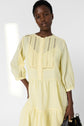 A person wearing a light yellow, long-sleeve, buttoned dress with lace and pleated details stands against a plain, light background, holding a black handbag.