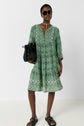 Woman in a green patterned dress featuring a motif géométrique, black sandals, and sunglasses stands against a white background, holding a black textured handbag and wearing a long necklace for an effortless style bohème.