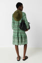 Person standing facing away wearing a green patterned dress with un motif géométrique, styled in a robe courte femme of style bohème, paired with a visor hat, sandals, and sunglasses, holding two black woven bags against a plain white background.