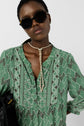 A person wearing large dark sunglasses and a green shirt with a motif géométrique and a rope necklace poses against a plain background, holding a black bag, showcasing an effortless style bohème.