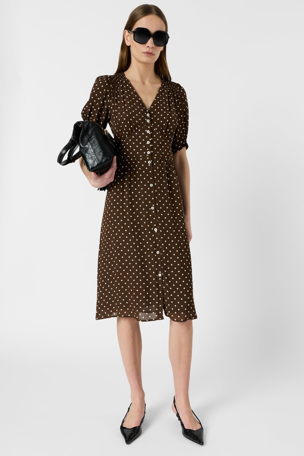 Woman in a brown, polka dot, button-down dress with puff sleeves, wearing black sunglasses and pointed shoes, holding a black handbag, standing against a plain white background.