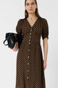 A model wearing a brown, V-neck midi dress with white polka dots, puff sleeves, and a button-down front, holding a black leather clutch bag against a plain white background.