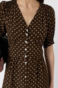 A person wearing a brown dress with white polka dots and buttons down the front, holding a black object under their arm. The dress has short sleeves and a V-neckline.