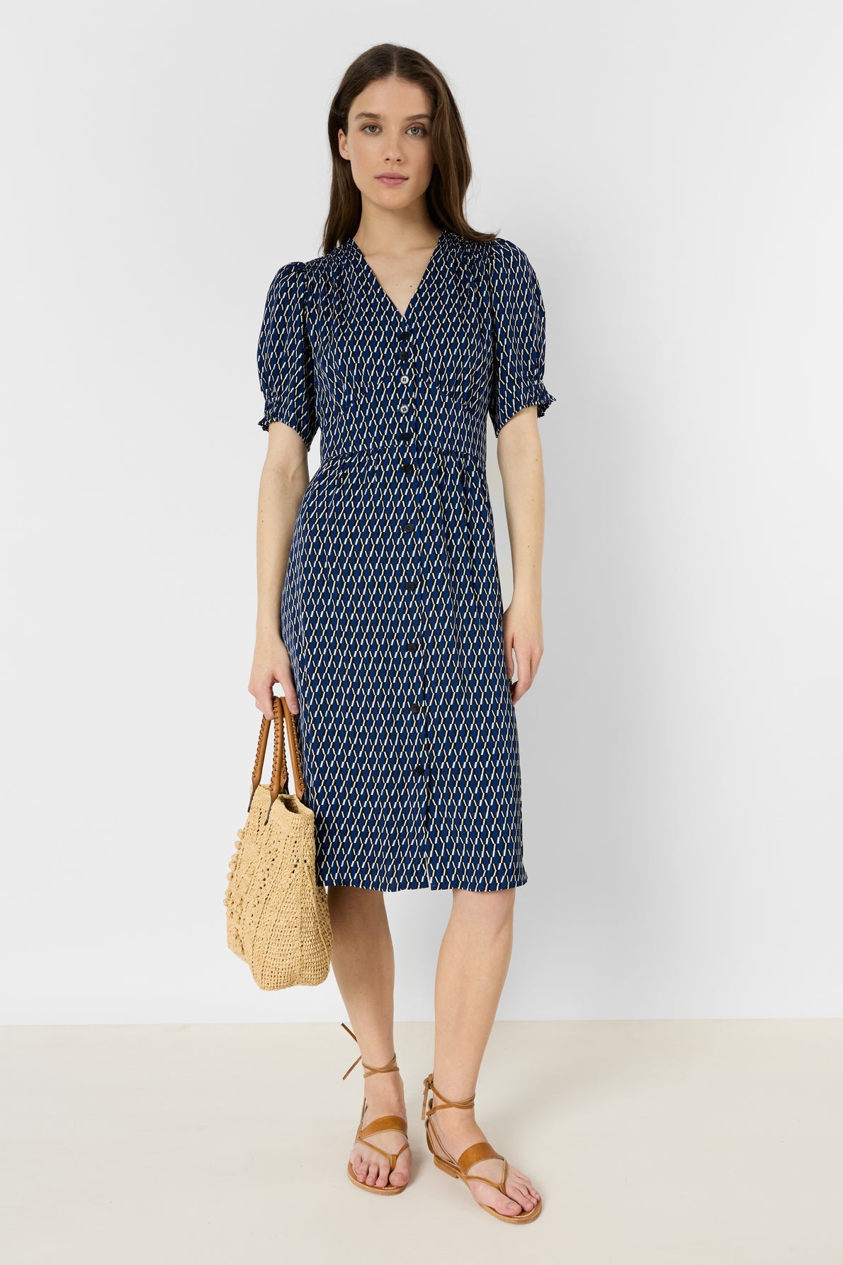 Woman standing against a white background, wearing a robe mi-longue bleu à imprimé graphique, tan sandals, and holding a woven beige handbag.
