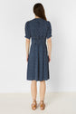 A woman stands facing away, wearing a robe mi-longue navy blue patterned dress with short sleeves and brown lace-up sandals. She has long brown hair and is in front of a plain white background.