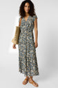 Woman with curly hair wearing a blue and yellow floral dress, tan sandals, and carrying a woven straw tote bag, standing against a plain white background.