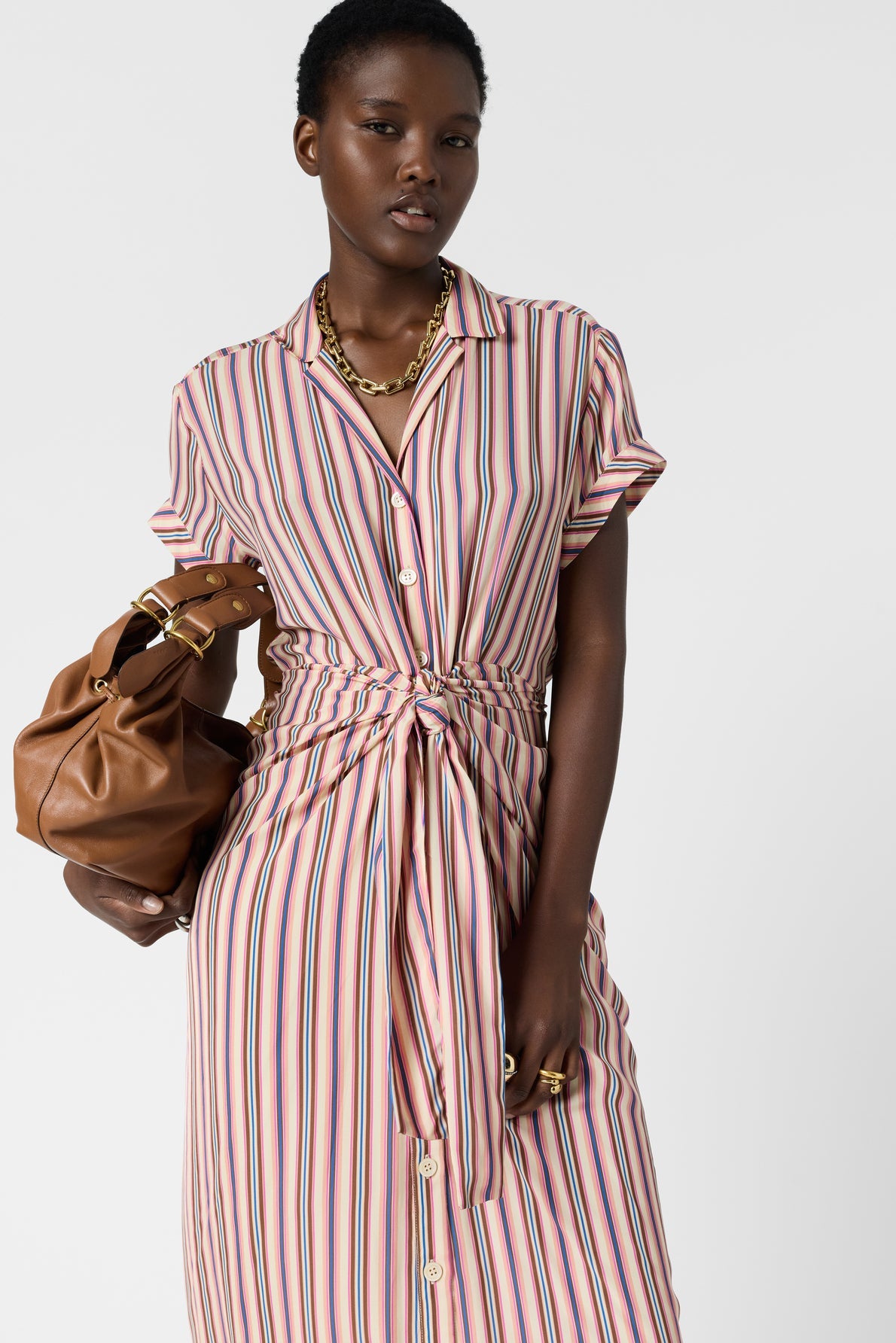 A person wearing a pink, blue, and beige robe chemise with rayures multicolores and a front tie holds a brown handbag. They have short hair, a gold chain necklace, and rings. The background is plain white.