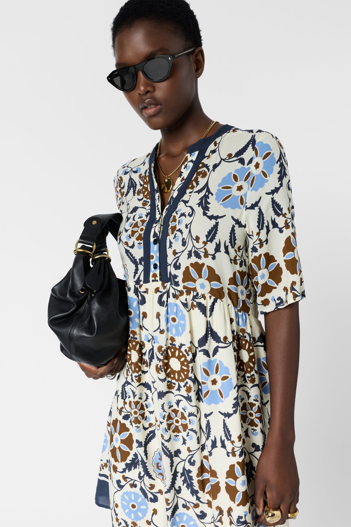 A person wearing a floral-patterned dress, dark sunglasses, and gold jewelry holds a black handbag. They have short hair and a confident expression, standing against a plain white background.