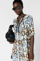 A person wearing a floral-patterned dress, dark sunglasses, and gold jewelry holds a black handbag. They have short hair and a confident expression, standing against a plain white background.