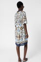 A person stands facing away, wearing sunglasses and a knee-length dress with blue, brown, and cream floral patterns, and black sandals, against a plain white background.