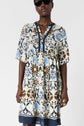 A person wearing sunglasses and a loose, patterned dress with blue, brown, and cream floral designs stands against a plain white background. The dress has short sleeves and a navy-trimmed V-neckline.