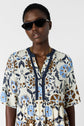 A person wearing black sunglasses, a gold necklace, and a floral-patterned, blue, brown, and cream dress stands against a plain light background.