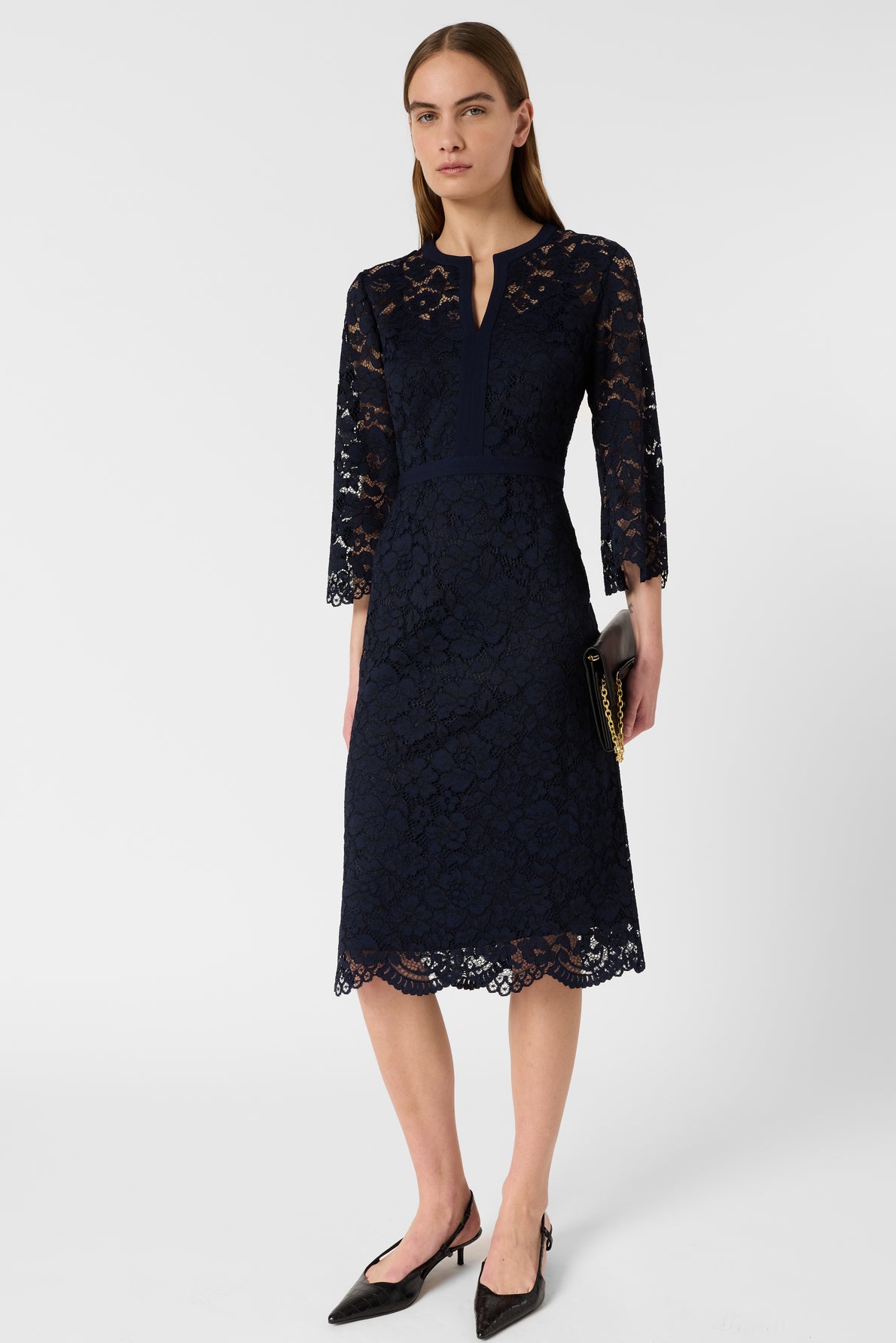 A woman stands against a plain background wearing a robe élégante bleu marine, crafted from delicate lace in a knee-length silhouette with three-quarter sleeves. She holds a black clutch and pairs the look with black pointed-toe heels.