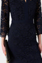 A person wearing a robe élégante bleu marine en dentelle with three-quarter sleeves holds a black clutch with a gold chain. The robe mi-longue dentelle features a floral pattern and a fitted waist.