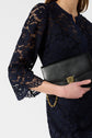 A woman in a robe bleu marine mi-longue dentelle holds a black clutch with a gold clasp and chain. Her face is partially out of frame, and the background is plain and white.