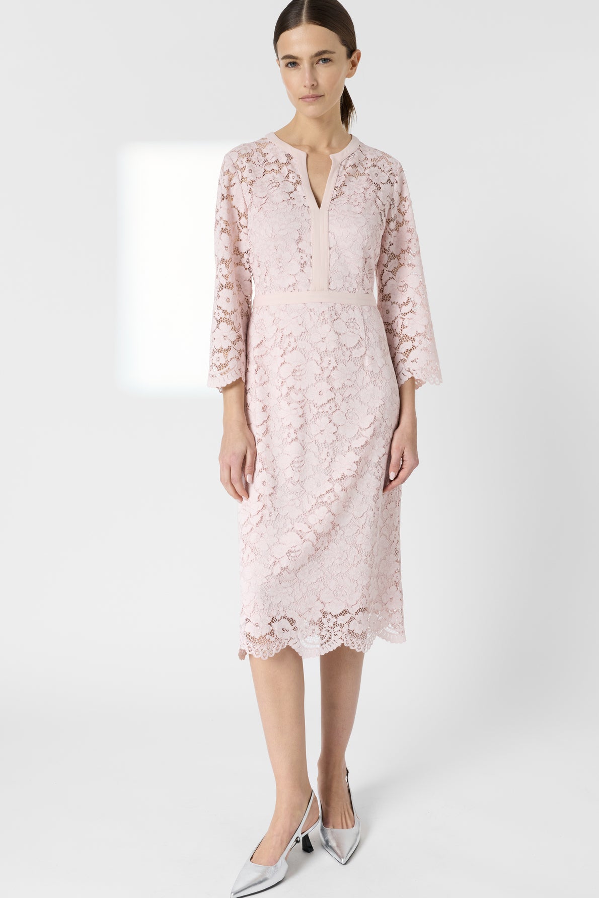 Woman wearing a pale pink, knee-length robe dentelle with three-quarter sleeves and a front keyhole detail, paired with silver pointed-toe heels, standing against a plain white background.
