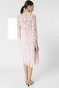 Woman wearing a pale pink, knee-length lace dress (robe cérémonie) with three-quarter sleeves and white open-toe heels, standing with her back to the camera against a plain light background.