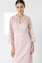 A woman wearing a light pink, robe dentelle with three-quarter sleeves stands against a plain white background. The mi-longue dress features a subtle floral pattern and a V-shaped neckline, making it an elegant choice for any cérémonie.