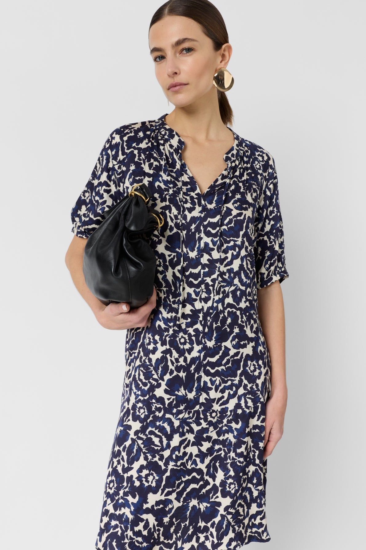 Woman with brown hair in a low ponytail wears a robe courte in blue and white motif floral, gold hoop earrings, and holds a black clutch bag under her arm, standing against a plain white background.