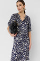 Woman with brown hair in a low ponytail wears a robe courte in blue and white motif floral, gold hoop earrings, and holds a black clutch bag under her arm, standing against a plain white background.