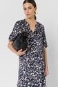 Woman with straight brown hair in a blue and white patterned robe courte holds a black handbag. She wears gold hoop earrings and stands against a plain, light background, showcasing the elegant motif floral in soft twill de viscose.