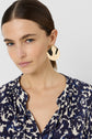 Woman with straight brown hair, wearing a navy and white patterned blouse reminiscent of a motif floral robe courte in twill de viscose, and large, abstract gold earrings, looking slightly to the side against a plain light background.