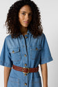A woman with curly hair wears a short-sleeved denim button-up dress with front pockets and a brown woven belt, standing against a plain light background.