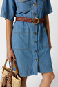 A person wearing a buttoned denim dress with a brown braided belt holds a straw handbag with a brown patterned scarf tied to the handle.