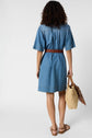 Woman with curly hair, wearing a knee-length denim dress with a brown braided belt and sandals, holding a woven straw bag with a brown scarf, standing and facing away from the camera.