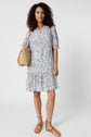 Woman with curly hair wears a robe courte fleurie bleu clair à col montant à volants, tan sandals, and holds a woven straw bag, standing against a plain white background.