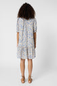 A woman with curly hair stands facing away, wearing a robe fleurie— a light blue and cream floral dress with short sleeves, col montant à volants, and a ruffled hem. She pairs it with tan lace-up sandals, set against a plain white background.