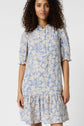 Woman wearing a robe courte bleu clair à motif fleuri beige, avec manches courtes bouffantes, col montant à volants légèrement fendu, et ourlet volanté, se tenant devant un fond blanc uni.