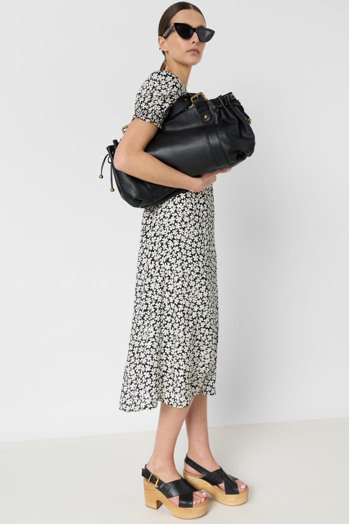 Woman in black cat-eye sunglasses, a black and white floral dress, and platform sandals holds a large black leather bag against a plain white background.