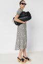 Woman in black cat-eye sunglasses, a black and white floral dress, and platform sandals holds a large black leather bag against a plain white background.