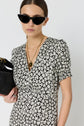 Woman wearing a black-and-white floral dress, black cat-eye sunglasses, and gold necklaces, holding a small black bag, standing against a plain white background.