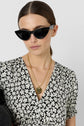 Woman with straight brown hair in a low ponytail wears black cat-eye sunglasses, a black and white floral dress, and layered gold necklaces with round pendants against a plain light background.