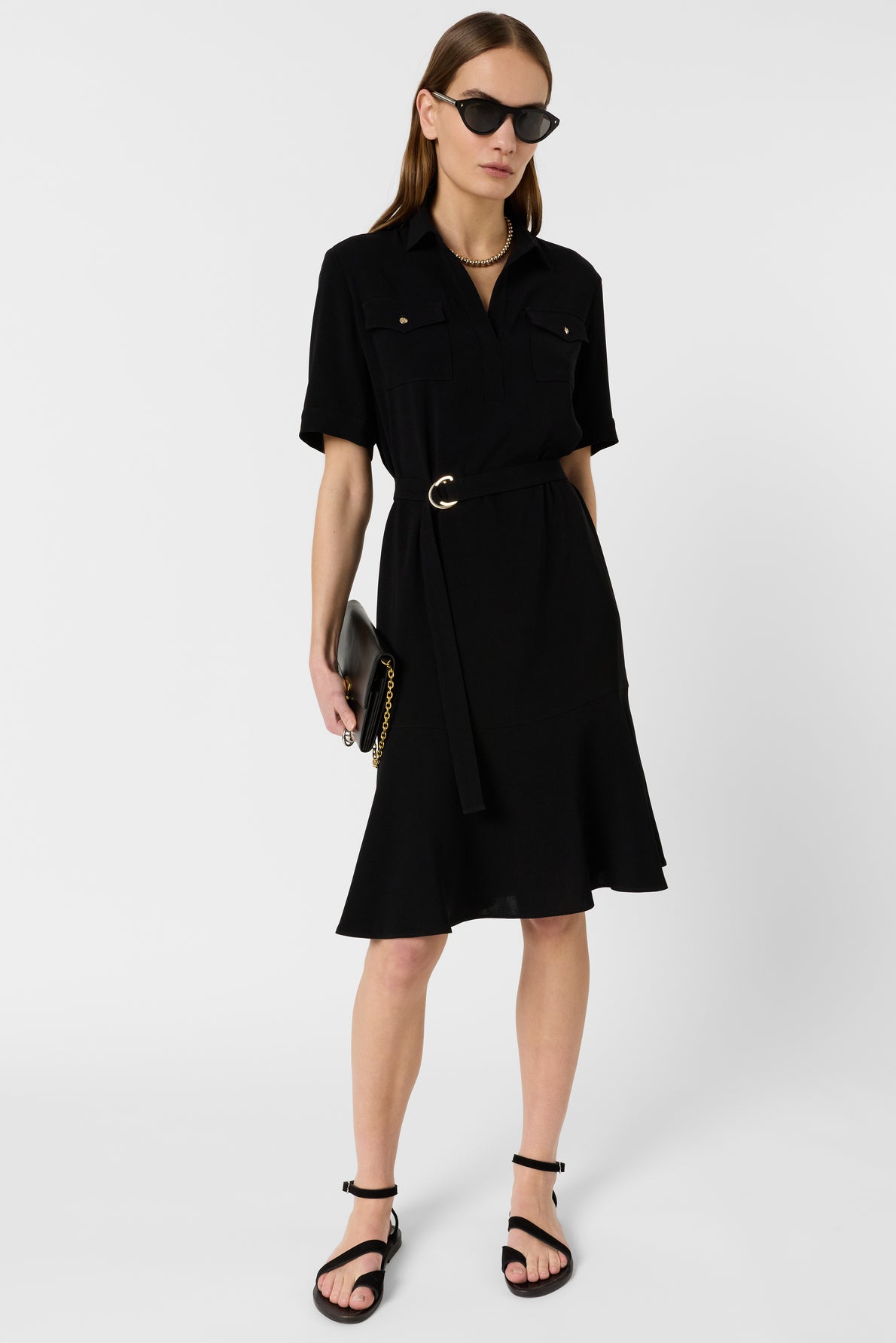 A woman wearing a black belted short-sleeve dress, black sandals, dark sunglasses, and holding a black clutch with a gold chain stands against a plain white background.