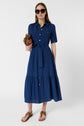 Woman in a blue, button-front, belted midi dress with short sleeves, brown sandals, large sunglasses, and holding a brown clutch bag, standing against a plain white background.