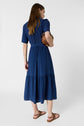 Woman standing and facing away, wearing a blue, short-sleeved, midi dress with a gathered waist and tiered skirt, holding a brown bag, and wearing brown sandals against a plain white background.