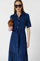 Woman wearing sunglasses and a blue belted button-down dress, holding a brown purse, standing against a plain white background.