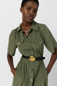 Person wearing a robe chemise kaki—an olive green button-up shirt dress with short sleeves, front pockets, a black belt with a gold buckle, and a chunky gold chain necklace—standing against a plain light background.