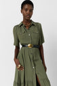 A person wearing a robe chemise kaki, styled as an olive green button-down dress with a black belt, gold buckle, chunky gold necklace, and rings, stands against a plain white background.