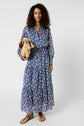 Woman with curly hair wearing a long blue floral dress, tan sandals, and holding a woven straw bag with a brown handle, standing against a plain white background.