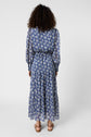 A person with curly hair, seen from behind, is wearing a long-sleeve, ankle-length blue floral dress and sandals, standing against a plain white background.